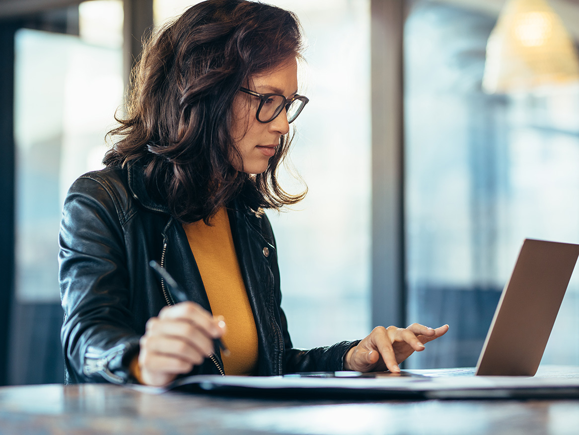 Businesswoman making notes looking at a laptop computer at office. Woman entrepreneur sitting at the table writing notes while working on laptop.
